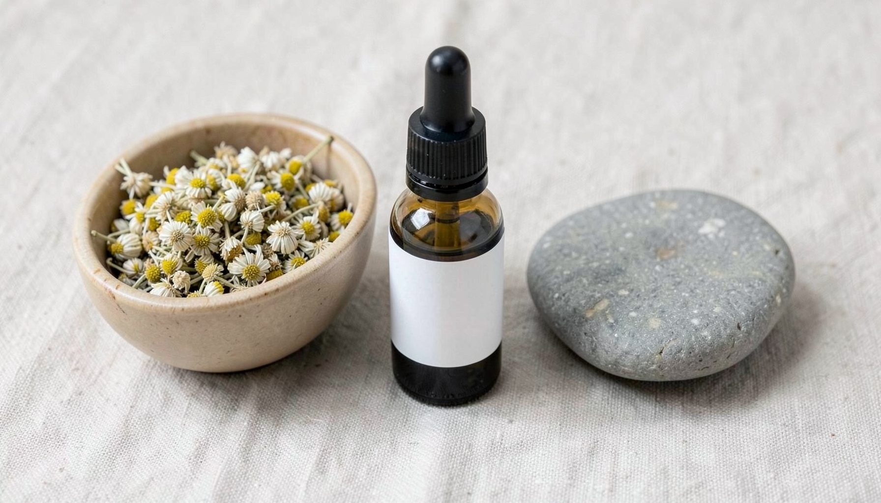 A small collection of naturopathy-inspired items: a glass dropper bottle, dried chamomile flowers in a bowl, and a smooth stone on linen