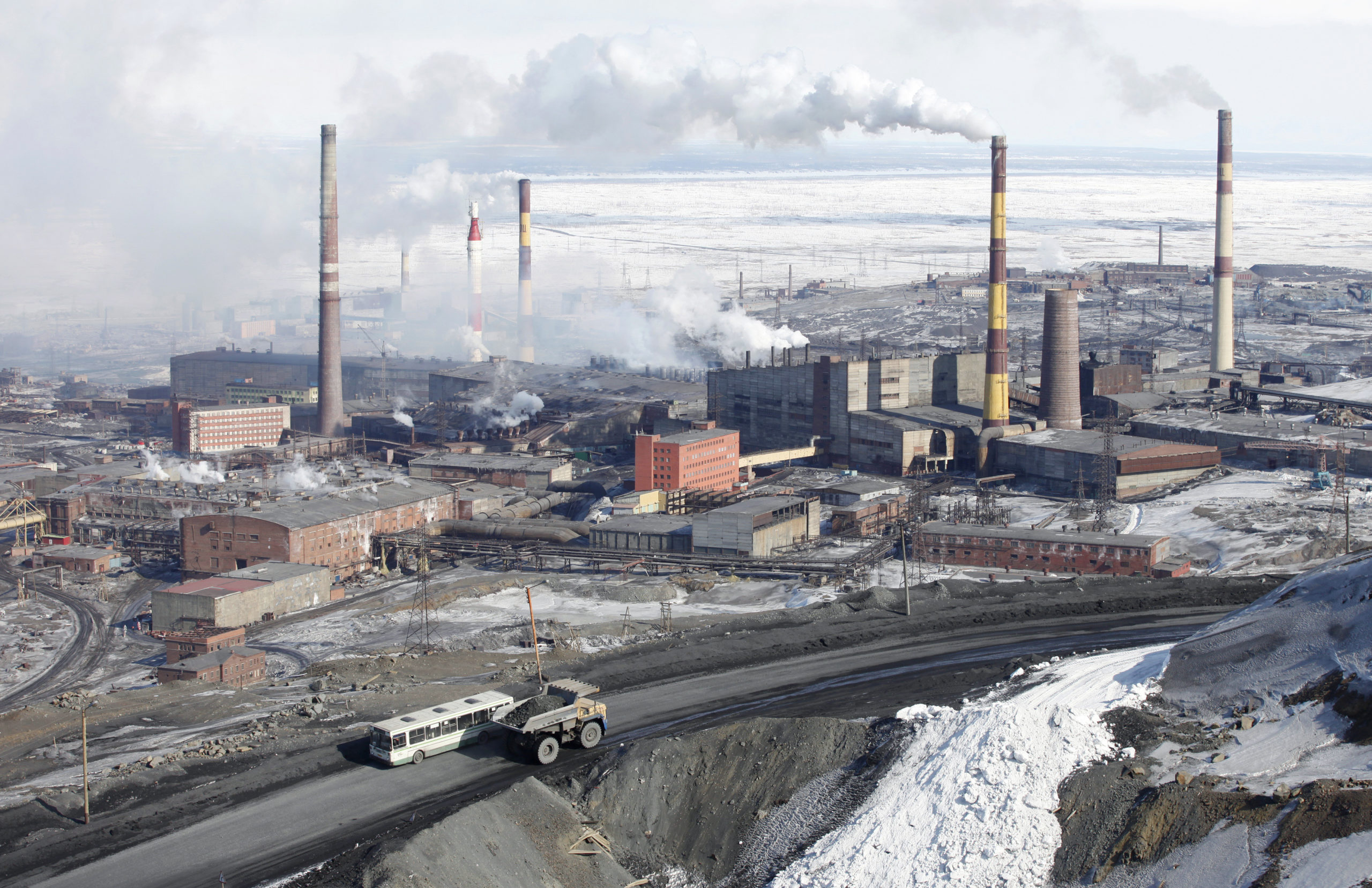 A serene winter landscape with an industrial complex in the distance, tall chimneys releasing soft plumes of smoke against a pale sky, snow covering the ground and rooftops, evoking a quiet, contemplative mood.