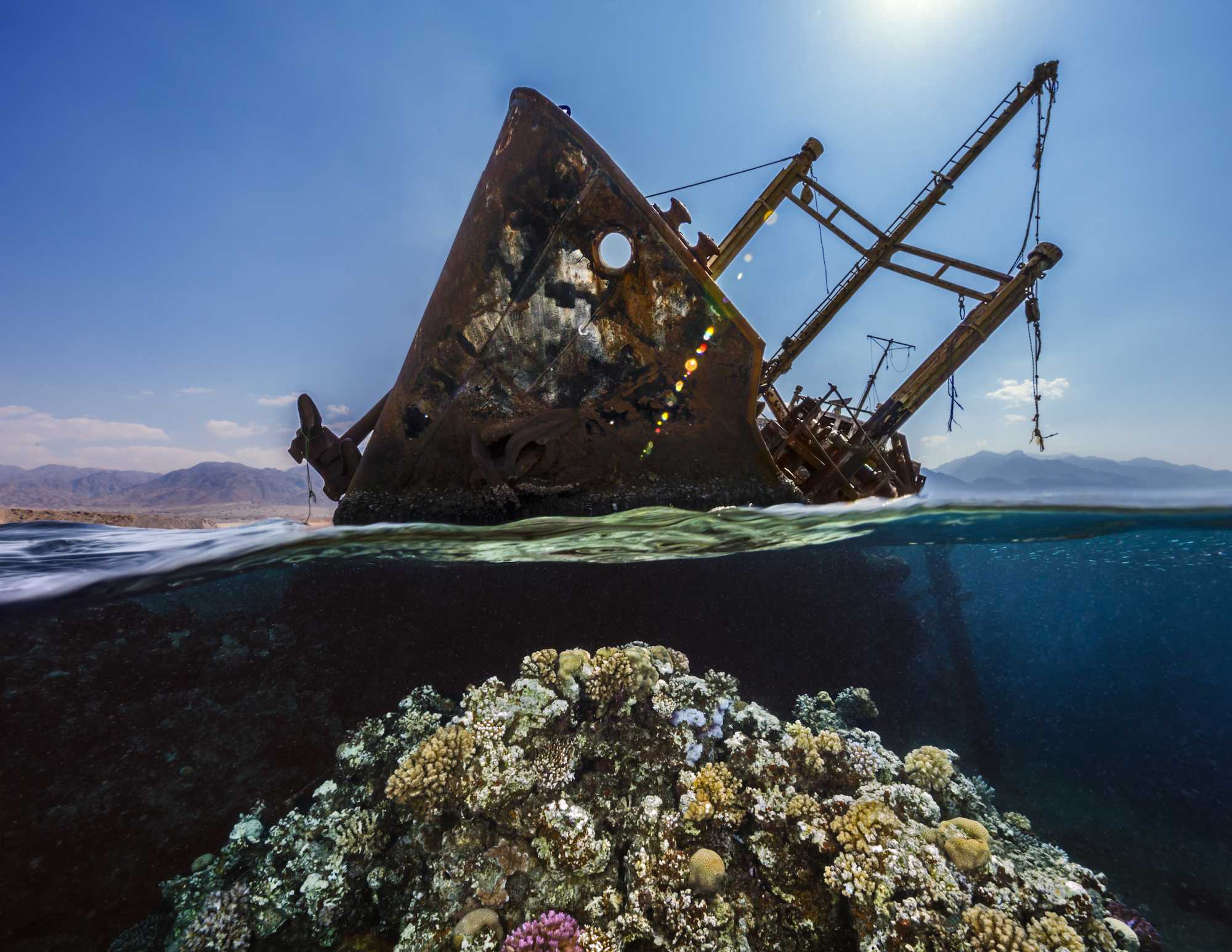 A split-view photograph of a rusted shipwreck above water and vibrant coral reef below, with distant mountains under a soft sky